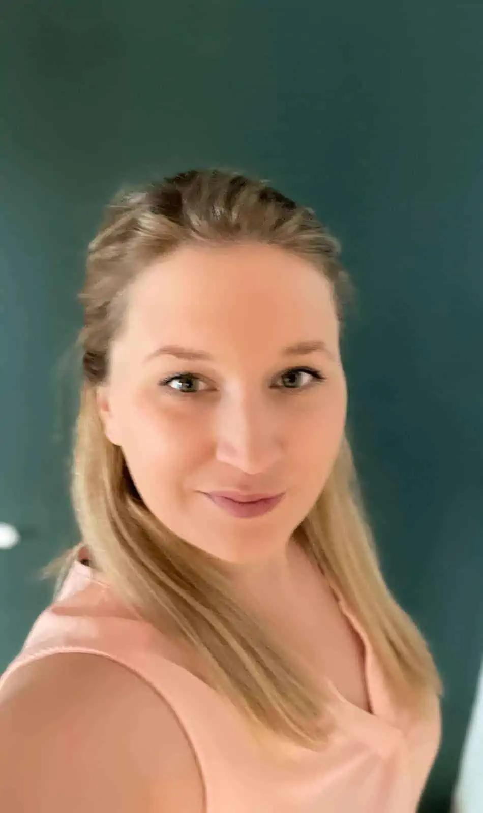 Eine lächelnde Person mit langen blonden Haaren trägt ein hellrosa Oberteil und steht vor einem dunkelgrünen Hintergrund.